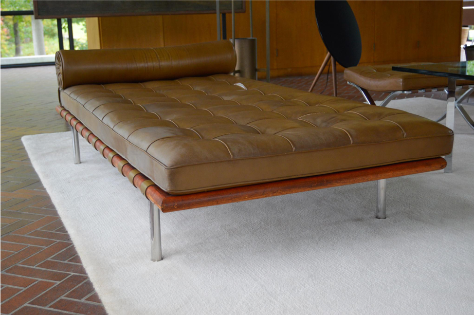 A photo of a couch in the living room of the Glass House. The brown-leather modern couch sits on a white carpet over a herringbone laid brick floor. 