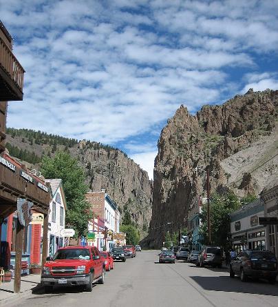Looking down the middle of the street. Cars are parked on either side and storefronts line the road. The stores melt into rocky cliff faces at the end of the street.