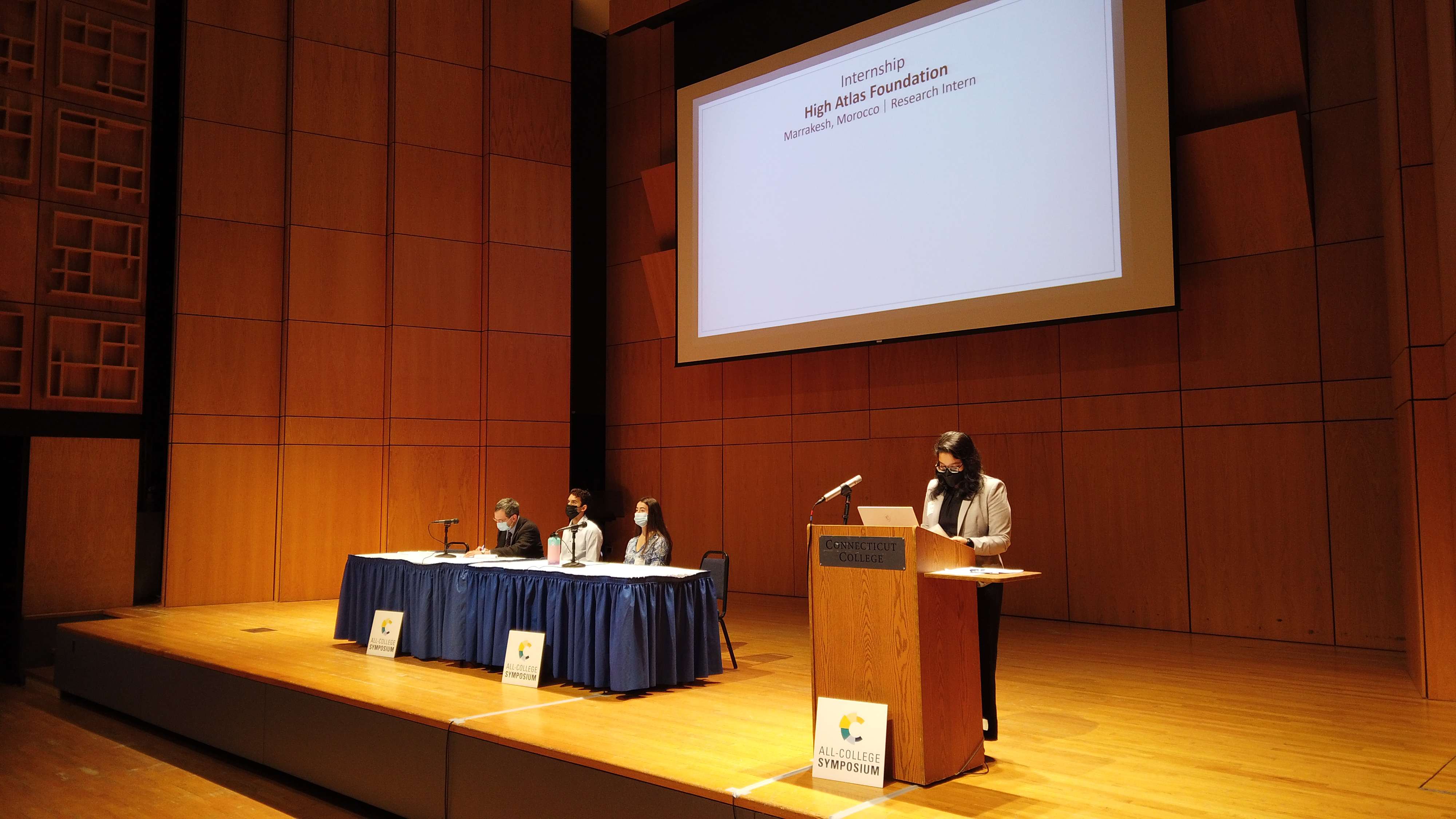Samirah stands behind the podium in front of a projector to present her project at the All-College Symposium. A panel of students sits on the stage awaiting their turn.